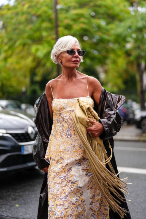 Grece Ghanem wears black sunglasses, a white and yellow off shoulders top with floral details, matching skirt, a dark brown leather jacket, a yellow...
