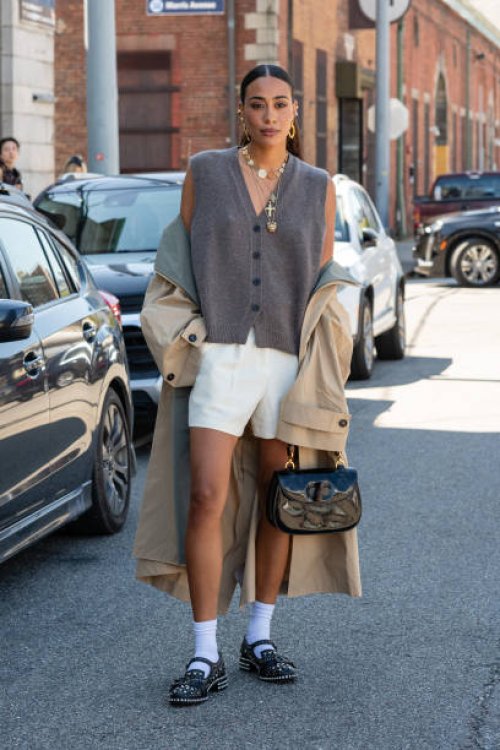Guest is seen wearing a grey vest, white shorts, tan trench coat, white socks, black shoes and carrying a black bag outside the COS show during New...