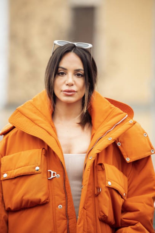 Guest is seen wearing white sunglasses, an orange oversized cargo parka with silver details, a white corset, white trousers, and orange velvet...