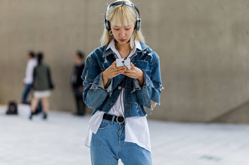 Guest wearing a ripped denim jacket and white button shirt searching for a music song on her phone while listening with her headphones on October 19,...