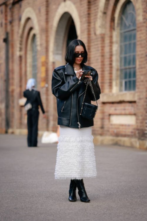 Guest wearing black oversized rider leather jacket, sunglasses and white knit dress and black boots during Australian Fashion Week Presented By...