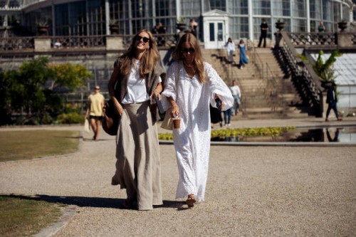 Guest wearing long grey skirt, white T-shirt, brown sweater over the shoulders, cup of ice coffee and big brown bag and a guest wearing long white...