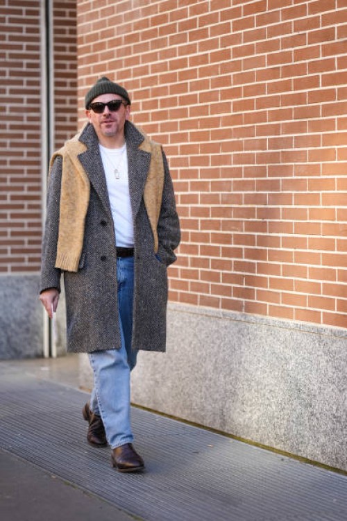 Guest wears a beanie hat, sunglasses, a beige scarf, a jacket, a white t-shirt, blue jeans, leather shoes, outside Fendi, during the Milan Fashion...