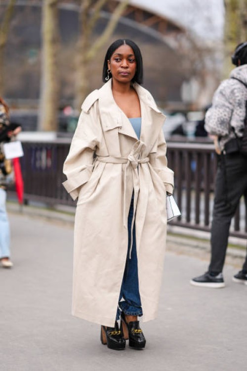 Guest wears a beige trench coat, a blue low neck top, a blue denim jeans / pants, black leather high heels shoes with golden chains, outside Chanel,...