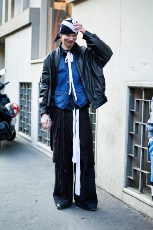 Guest wears a black and white printed bandana, a white lace as necklace, a black shirt, a denim gilet, a black leather jacket and black wide leg...