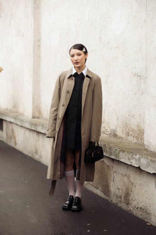 Guest wears a black dress, long white socks, black loafers, a brown trench coat, a black bag, and a metal Prada hair clip outside Prada during the...