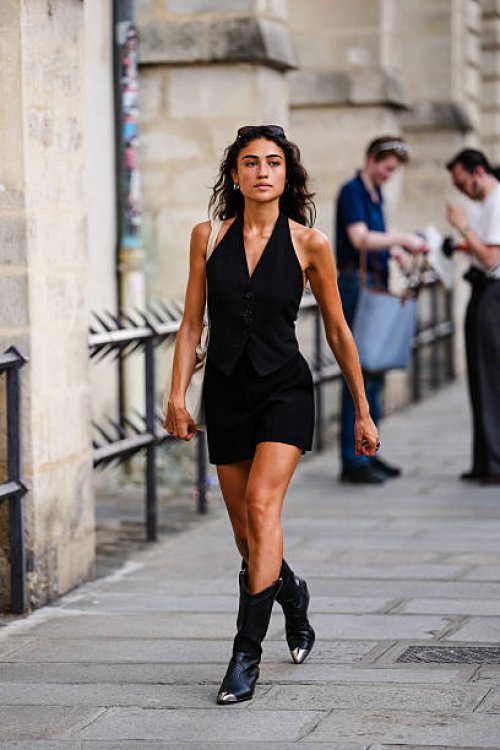Guest wears a black halter-neck vest with button detailing, paired with matching black shorts. The ensemble is complemented by black cowboy boots...