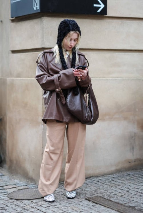 Guest wears a black hat with braids, a brown oversized leather jacket , a bag, beige flared pants, black and white shoes with crocodile patterns,...