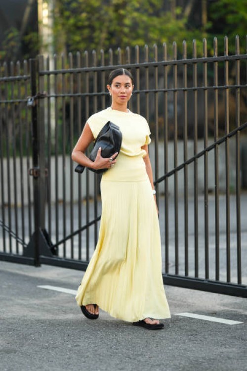Guest wears a black leather bag, earrings, a yellow sleeveless top, a pale pastel yellow pleated long skirt, sandals, outside COS, during New York...