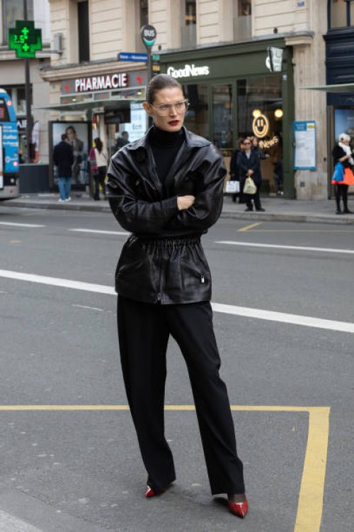 Guest wears a black leather jacket, black turtleneck, wide-leg black trousers, red pointed-toe heels, and clear-framed glasses outside Magda Butrym...