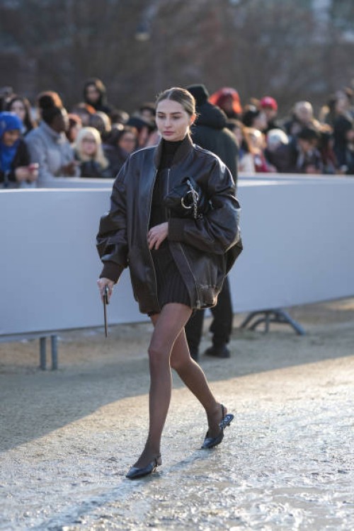 Guest wears a black leather oversize bomber jacket, a turtleneck black ribbed wool mini skirt, tights, leather shoes, outside Dior, during the...