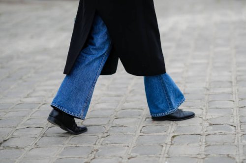 Guest wears a black long coat, blue denim flared pants, black shiny leather ankle shoes, outside Chanel, during Paris Fashion Week - Haute Couture...