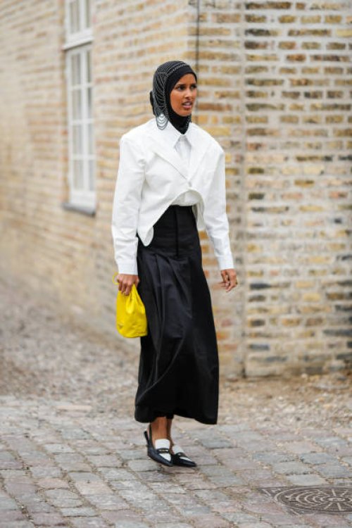 Guest wears a black scarf as a headband, a silver chain hat, a white asymmetric cropped blazer jacket, a white shirt, a black high waist long skirt,...