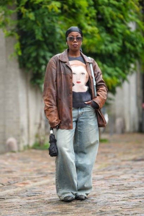 Guest wears a black silk scarf as a headband, black sunglasses, a black with beige face print pattern t-shirt, a dark brown shiny leather zipper...