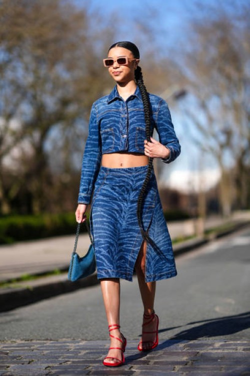 Guest wears a blue denim jacket as a cropped top, matching midi skirt with a slit, a blue bag, red high heels, sunglasses, outside Stella McCartney ,...