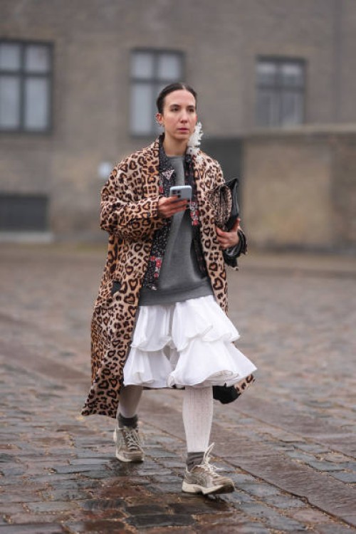 Guest wears a brow leopard print pattern long coat, a gray pullover, a white mini ruffled dress, sneakers shoes, outside Rolf Ekroth, during the...