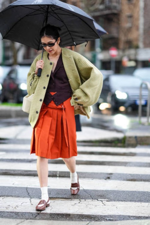 Guest wears a burgundy waistcoat, a green checked oversized jacket, sunglasses, a white leather bag, an orange midi pleated skirt, brown leather...