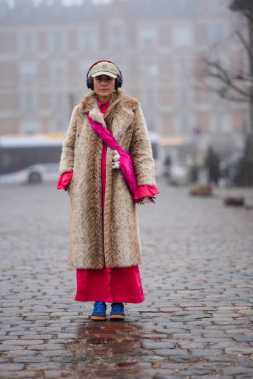 Guest wears a cap, a brown fluffy faux fur long coat, a pink dress, blue sneakers, headphones, outside Rolf Ekroth, during the Copenhagen Fashion...