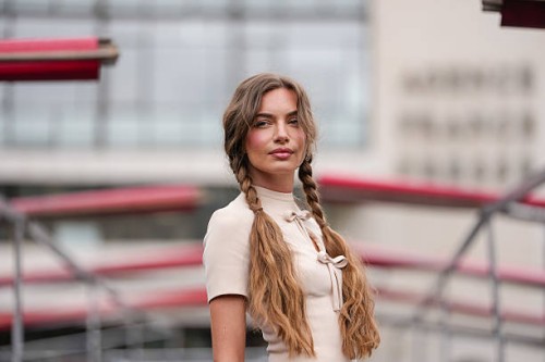 Guest wears a cream-colored, short-sleeved top featuring a high collar and a keyhole cutout with a bow detail at the front. Hair is styled in two...
