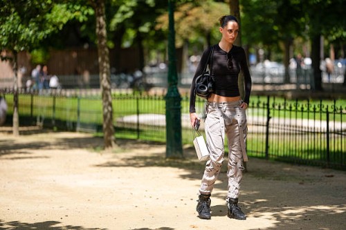 Guest wears a fitted, long-sleeve black top with a round neckline. Sunglasses are tucked into the neckline. Hair is styled in a sleek bun. Carries a...