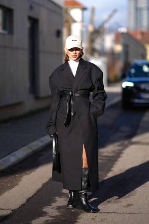Guest wears a gray cap hat, a white turtleneck pullover , a black long winter coat, a belt, knee high black leather boots , a leather bag, outside...