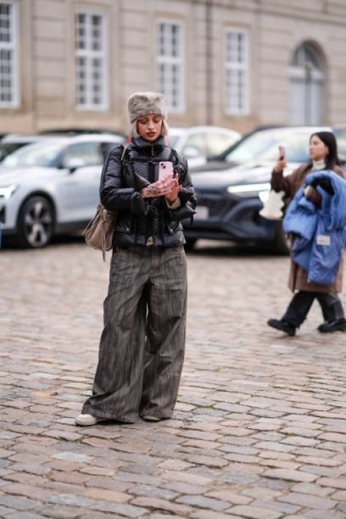 Guest wears a gray fluffy chapka hat, a black puffer jacket, flared pants , beige shoes, holds a mobile phone to take a picture / selfie, outside...