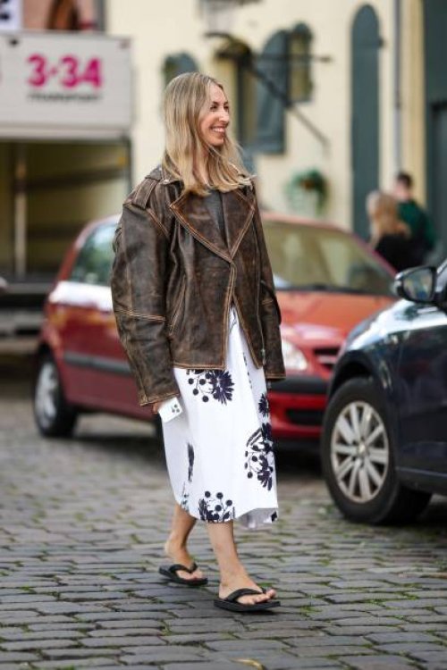 Guest wears a gray wool pullover, a dark brown faded denim oversized leather jacket, a white with embroidered black flower pattern midi dress, black...