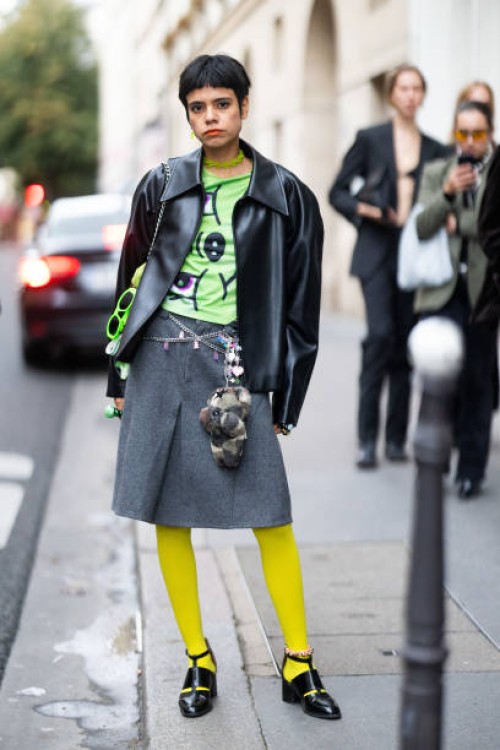 Guest wears a green necklace, a green printed t-shirt, a black leather jacket, a silver belt, a grey skirt, fluo green sunglasses, yellow tights and...