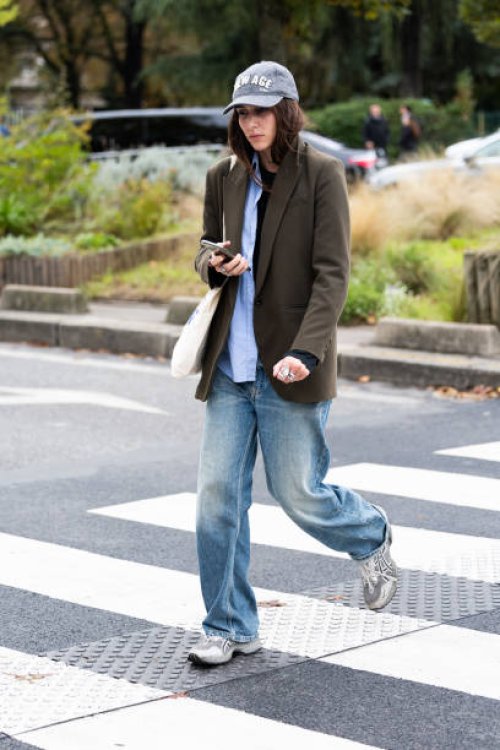 Guest wears a grey hat, a black t-shirt, a light blue shirt, a military green jacket, blue jeans, grey sneakers and a white tote bag outside Chloé...