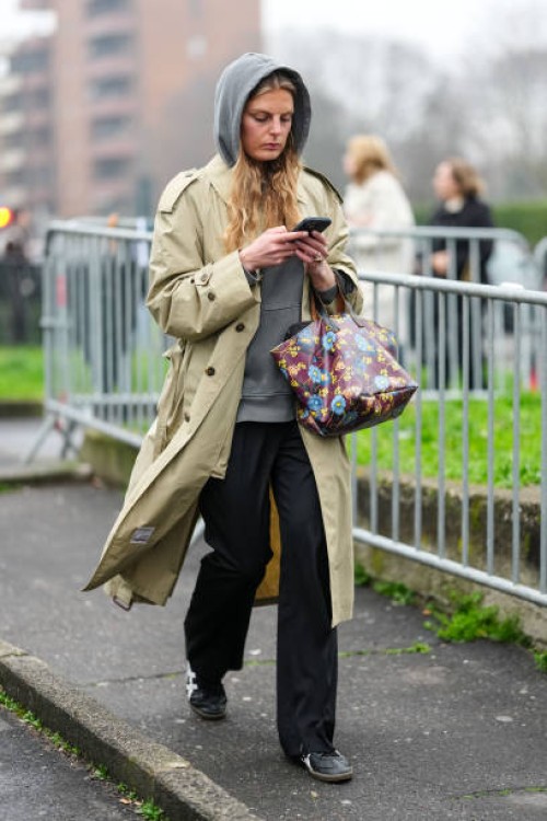 Guest wears a grey hoodie, a cream coat, black pants, burgundy leather bag with floral details, black leather sneakers shoes, outside Gucci, during...