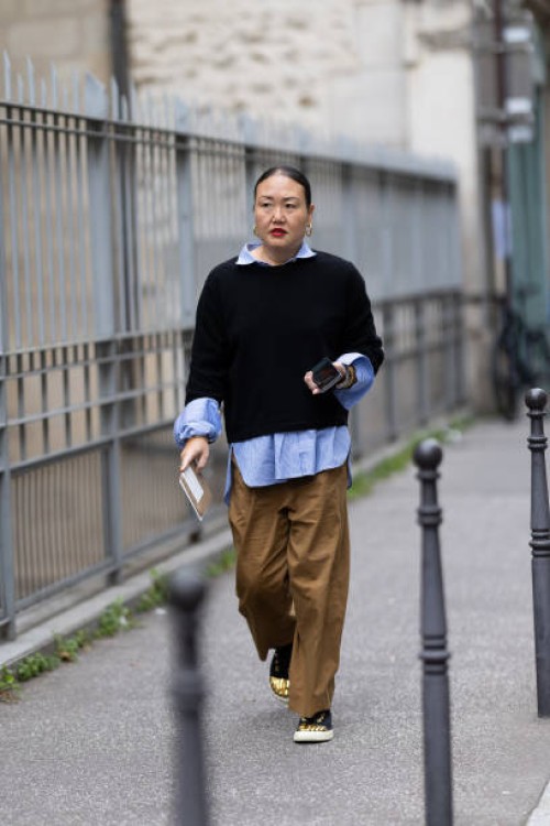Guest wears a light blue shirt, a blue sweater, beige trousers and black and gold sneakers outside Akris show during Womenswear Spring/Summer 2025 as...