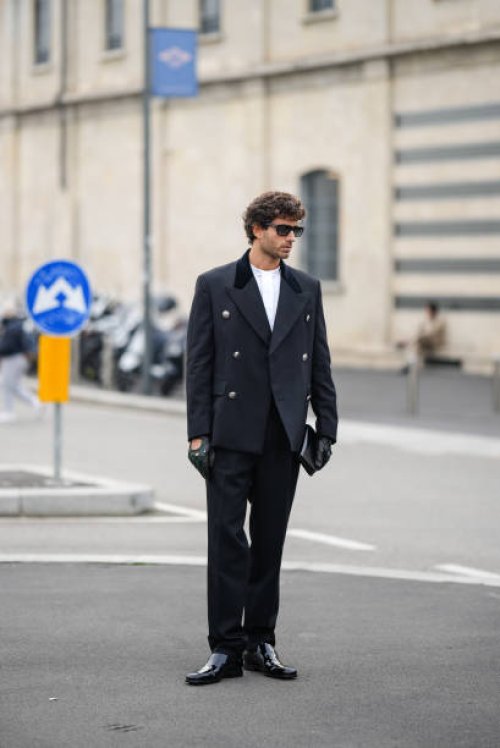Guest wears a military / blazer jacket, a white t-shirt, black leather gloves, black pants, black shiny leather pointed shoes, outside Prada, during...