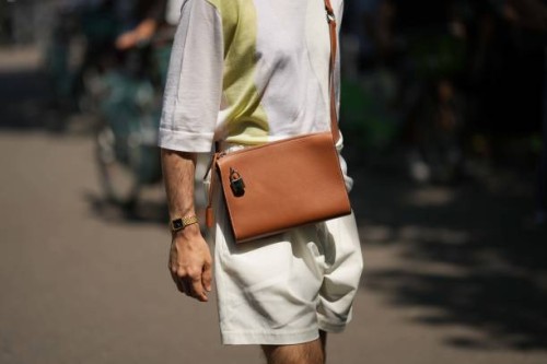 Guest wears a pale gray with large pale khaki strap pattern t-shirt, a camel shiny leather crossbody bag, beige suit shorts, a gold watch , outside...