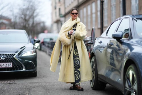 Guest wears a pastel pale yellow full look, an oversized scarf, sunglasses, a black and yellow dress with printed geometric patterns, brown leather...