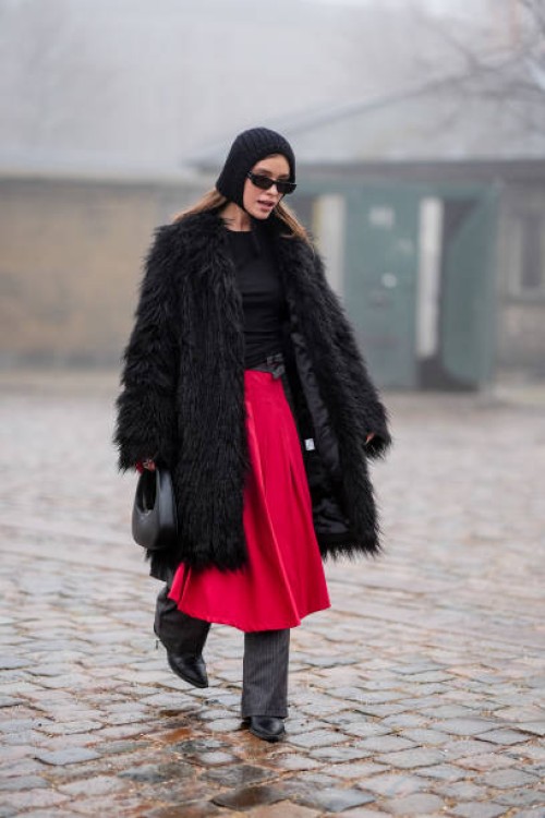 Guest wears a ribbed wool hood sunglasses, a black fluffy long coat, a black leather Coperni bag, a red skirt, gray striped flared pants, black...