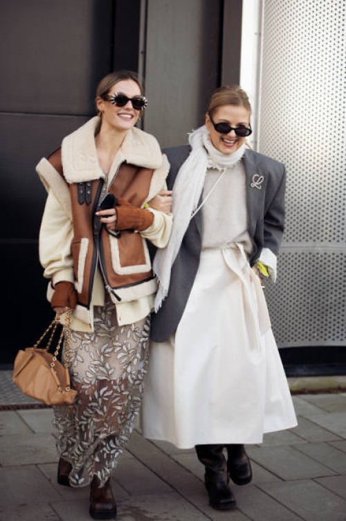 Guest wears a sheer long coat, white sweater, brown fur and leather vest, brown bag, and brown boots and a guest wears a white skirt, white sweater,...