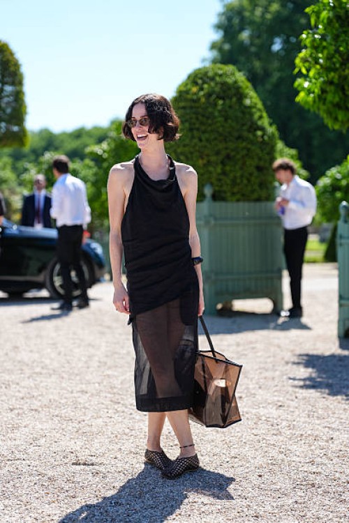 Guest wears a sleeveless black dress with a draped neckline and a sheer overlay extending below the knee. The hair is styled in a short, wavy bob....