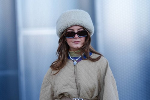 Guest wears a white fluffy chapka hat, sunglasses, a necklace, a green pullover , a beige padded oversized jacket, outside Munthe, during the...