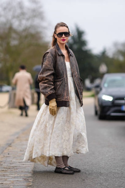 Guest wears a white gathered dress with low-neck and floral embroidery, a brown oversized leather jacket, beige gloves, sunglasses, black ballet...