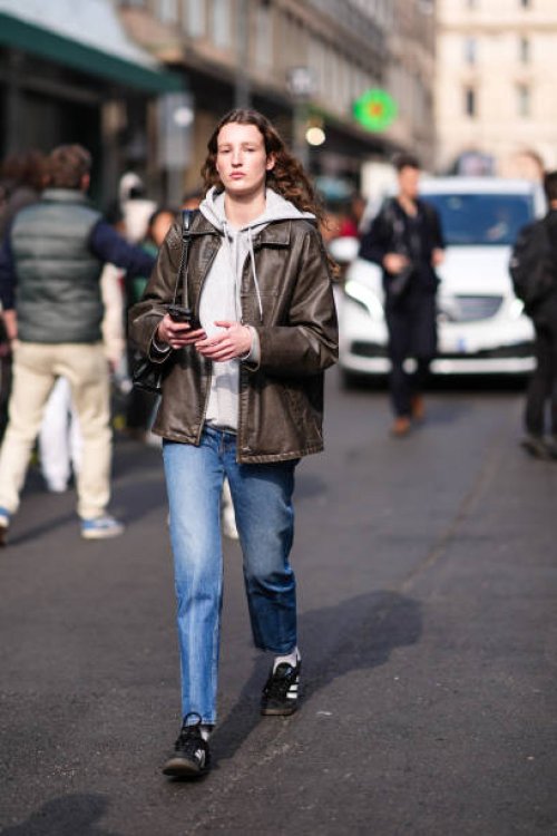 Guest wears a white / grey hoodie, a brown leather jacket, blue denim jeans / pants, a black leather bag, black and white sneakers, outside Onitsuka...