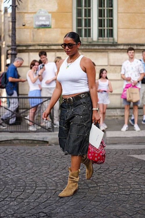 Guest wears a white ribbed tank top paired with dark denim knee-length shorts featuring prominent stitching and metal embellishments. Accessories...