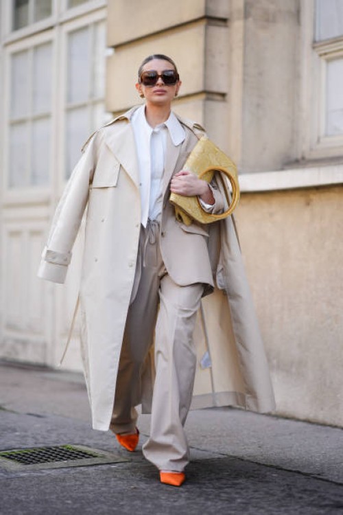 Guest wears a white shirt, a beige trench coat, beige wide-leg pants, orange pointed shoes, a yellow leather bag, sunglasses, outside Giambattista...