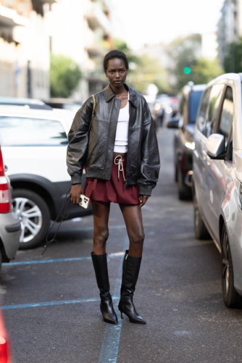 Guest wears a white tank top, a red burgundy mini puffy skirt, black leather boots and a brown leather jacket outside Max Mara show during the Milan...