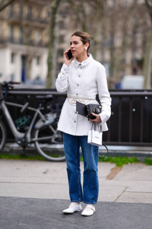 Guest wears a white tweed coat, a flower shaped earring, a black Chanel bag, blue denim jeans / pants, white ballerina shoes outside Chanel, during...