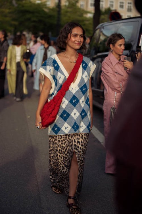 Guest wears animal print long skirt with split, white and blue BP checkered vest and red shoulder bag outside the Baum und Pferdgarten show during...