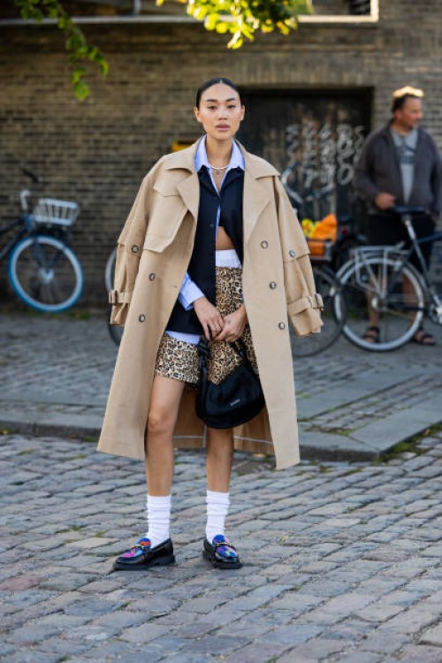 Guest wears animal print shorts, beige trench coat, blue button shirt, white socks, loafers outside Ganni during the Copenhagen Fashion Week...