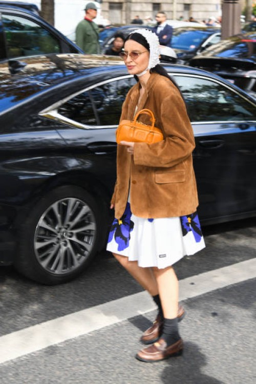 Guest wears beige Miu Miu top, white Miu Miu floral print skirt, brown suade blazer, orange Miu Miu bag, grey socks, brown loafers, outside Miu Miu...