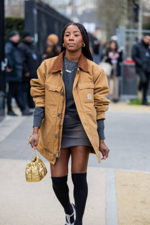 Guest wears beige oversized Carhartt jacket, grey jumper, skirt, golden bag, over knee high socks outside Lacoste during the Womenswear Fall/Winter...