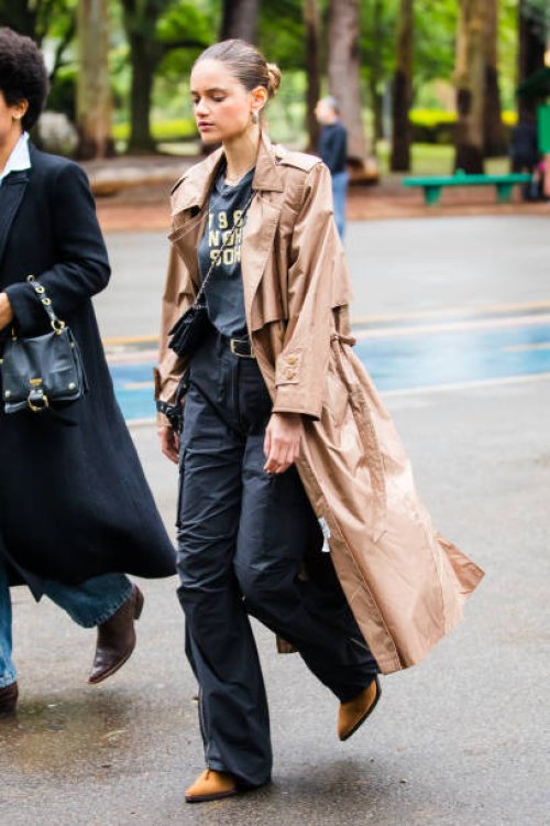 Guest wears beige trench coat, graphic t-shirt, black cargo pants, tan boots, small black crossbody bag, hoop earrings, slicked-back hair at Sao...