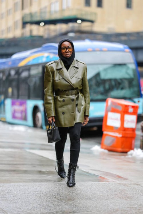 Guest wears black balaclava, black sunglasses with orange lenses, shiny olive green belted double breasted leather coat from Prada, black mini...
